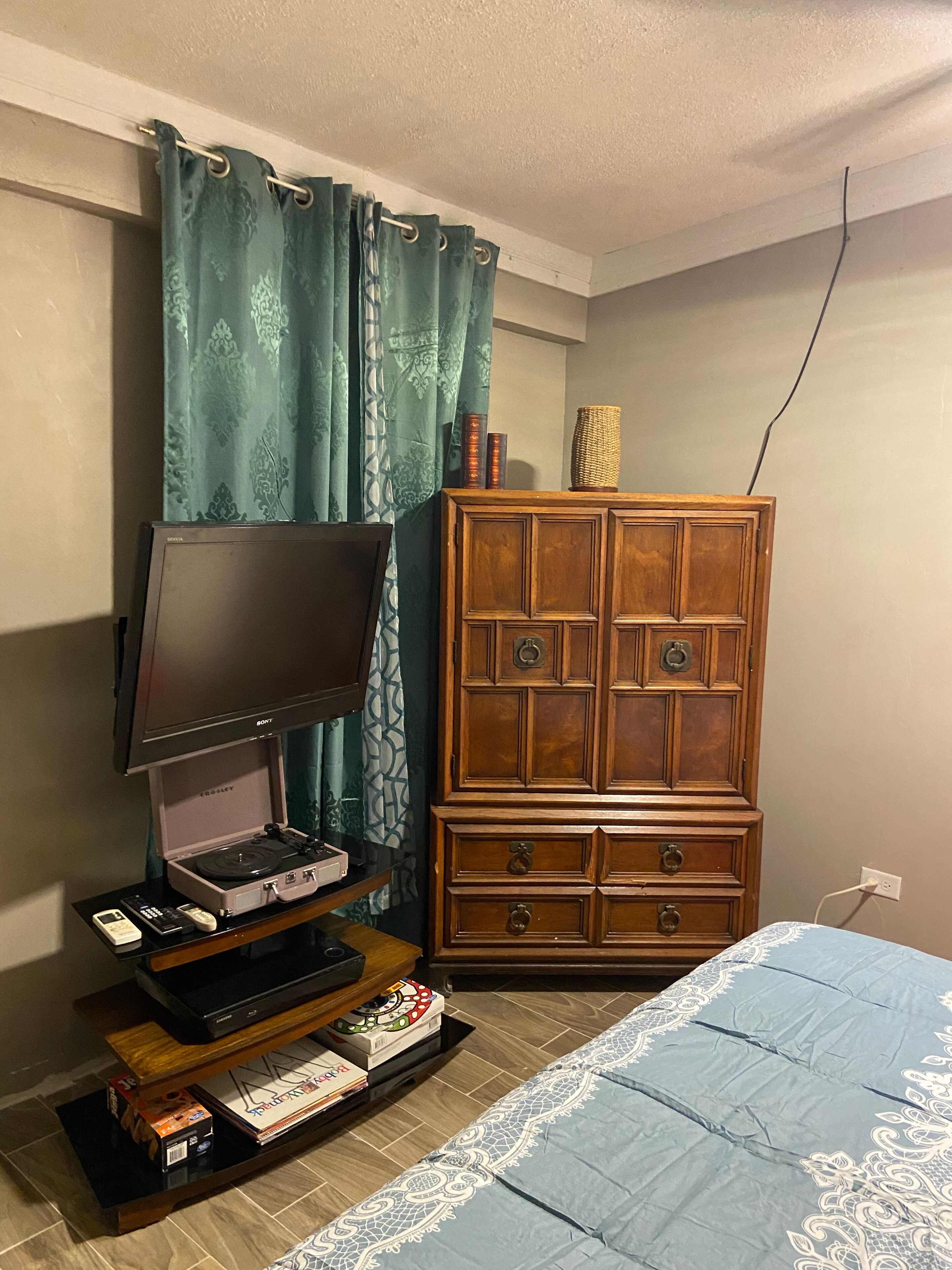 Una habitación con una televisión de pantalla plana sobre un pedestal, un armario de madera, una cama con colcha estampada y una cortina con diseño geométrico.