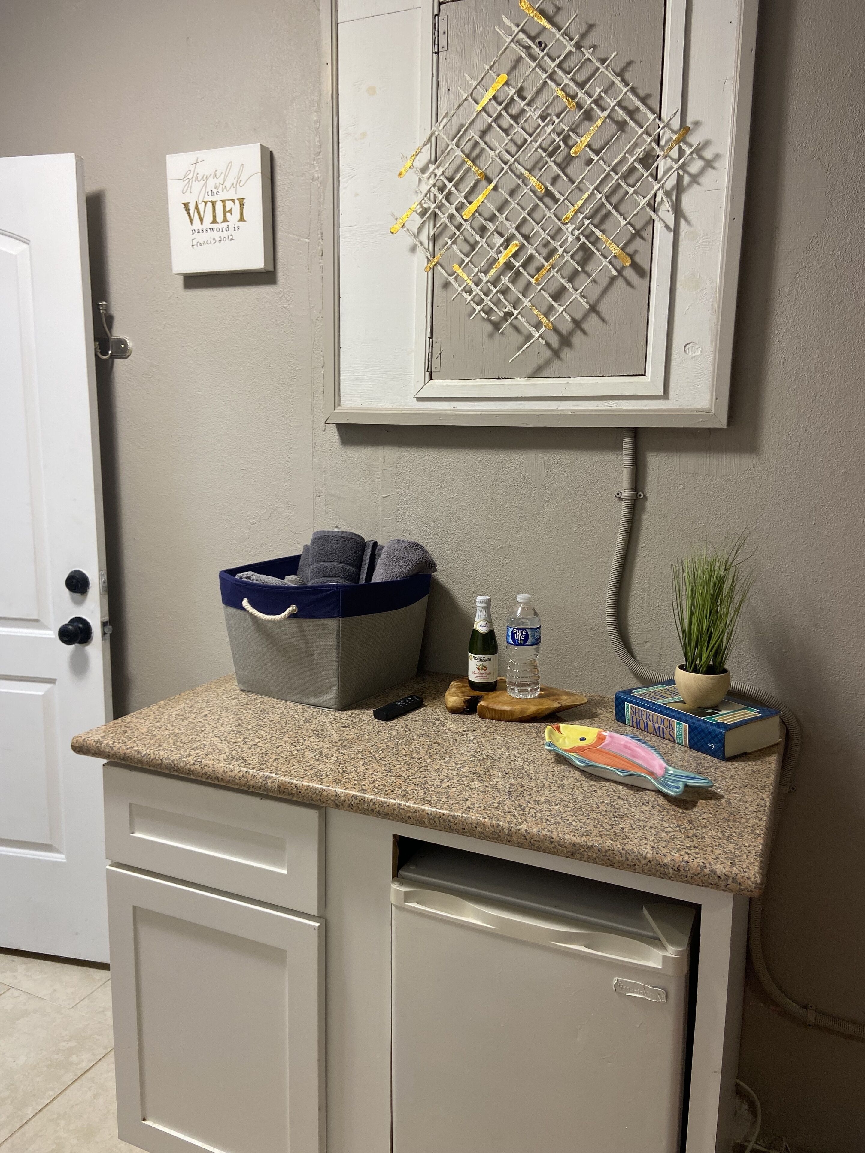 A kitchen counter with a blue basket, a remote control, a bottle, a water bottle, a book, and a fish decoration.