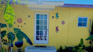 A yellow house with a white door, a small window, and a concrete pathway leading to the door. There are decorative lizards on the wall and a large plant to the left. - Rosebud (Rock Sound)