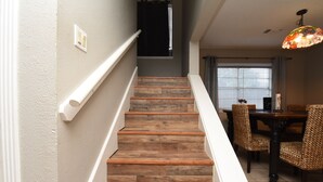 Un escalier avec des marches en bois et des rampes blanches, menant à un coin repas agrémenté de chaises en osier et d’une table.