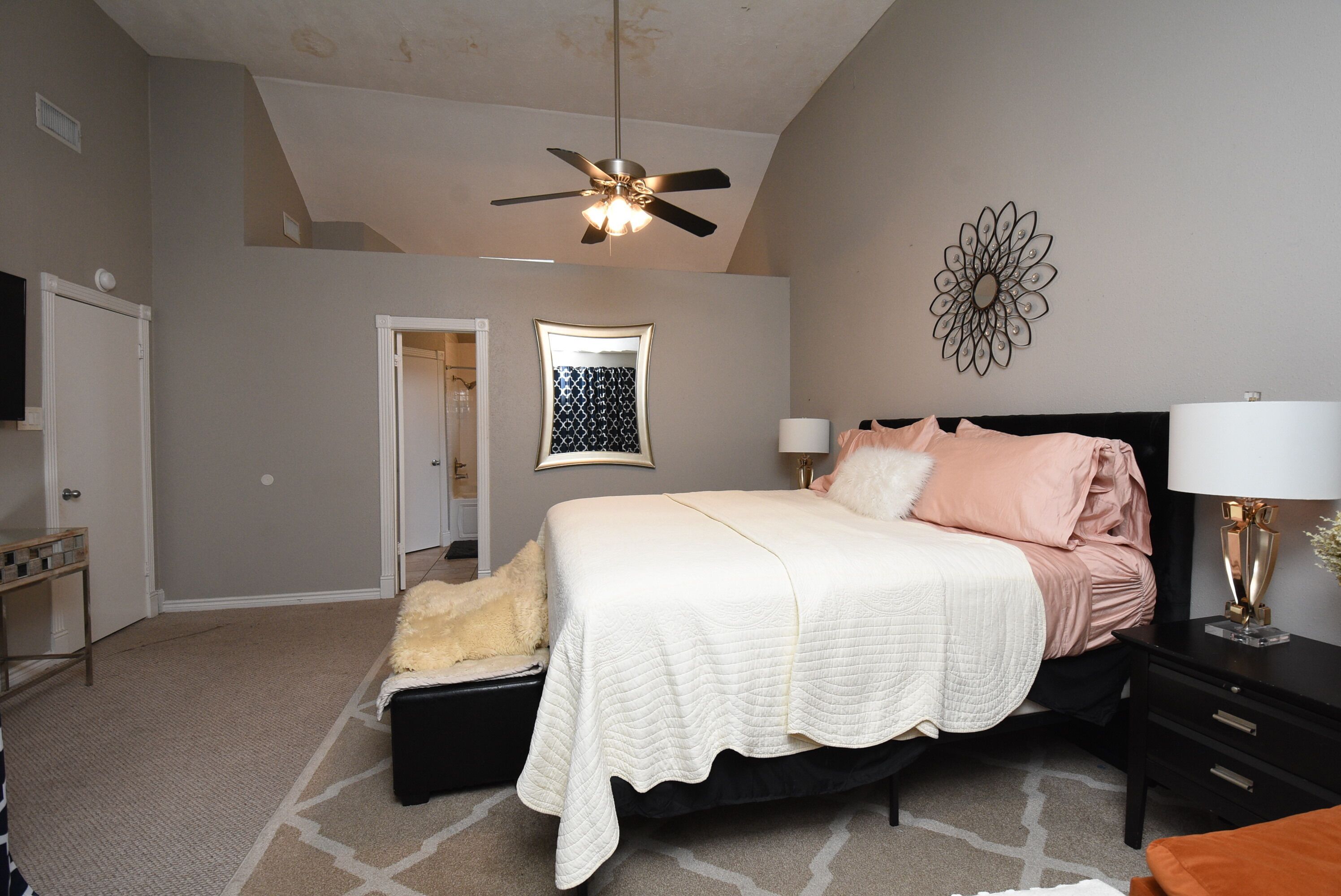 Une chambre à coucher comprenant un lit, un ventilateur de plafond, un luminaire fixé au mur, un élément décoratif mural et une table de chevet avec une lampe.