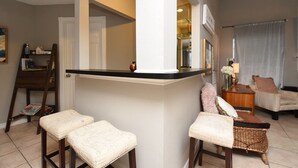 Un salon comprenant un coin cuisine, un banc en bois, une chaise en osier et un canapé blanc.