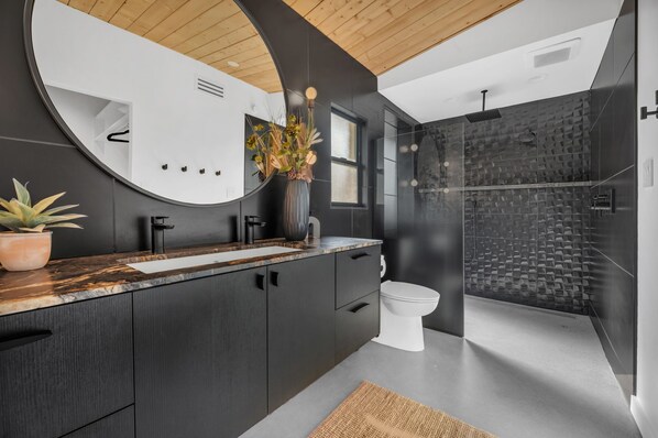 A modern bathroom with a large mirror, dark cabinetry, a white toilet, and a shower area with a textured wall.