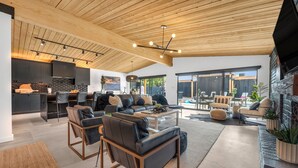 Uma sala de estar moderna com teto de madeira, sofás de couro preto, uma mesa de centro e uma área de cozinha com armários escuros e painel de pedra.