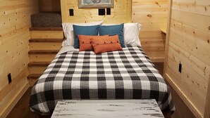 A cozy wooden room with a checkered bedspread, a painting on the wall, and a small table with a yellow object on it. - The Redbud Cabin at Roman Nose (Watonga)