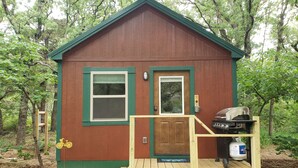 A small wooden cabin with a front door, a window, and a deck with a grill and propane tank. - The Redbud Cabin at Roman Nose (Watonga)