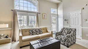 Un salon comprenant un canapé, un fauteuil et un pouf, de grandes fenêtres et une porte.