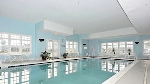 Une piscine intérieure à l’eau d’un bleu cristallin, entourée de chaises et de tables blanches, de grandes fenêtres et de plantes en pot.