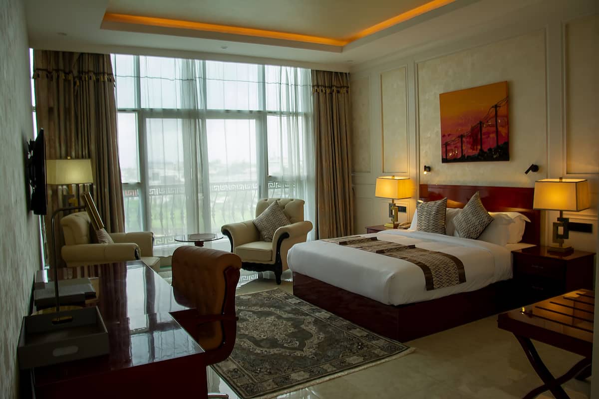 A hotel room with a large bed, two armchairs, a desk, and a view of the city through large windows.