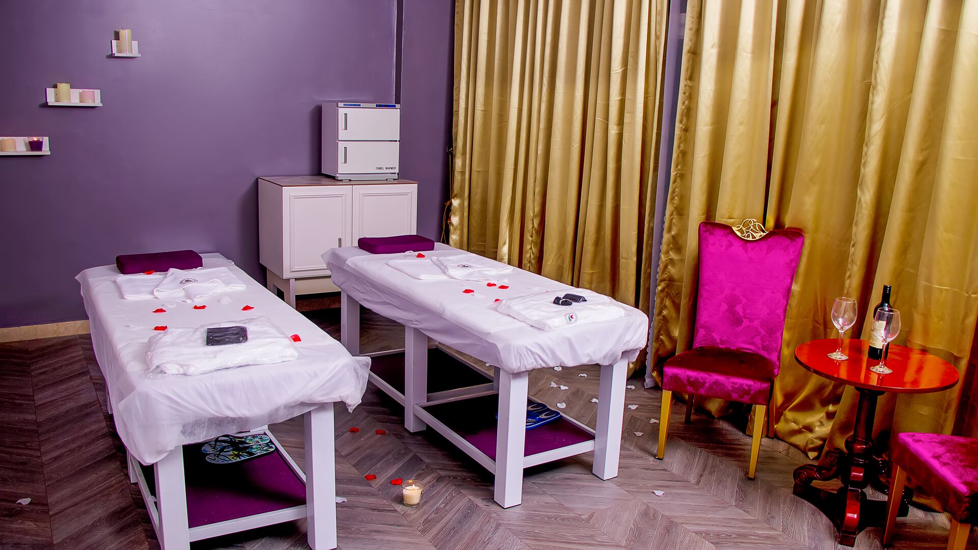 A room with two beds, a massage table, a red chair, a small table with a bottle and glasses, and a minibar.