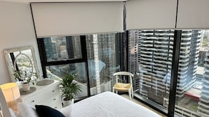 A modern bedroom with a large bed, a dresser, a chair, and a view of a cityscape through large windows. - 3 bedroom apartment in the heart of the CBD. (Melbourne)