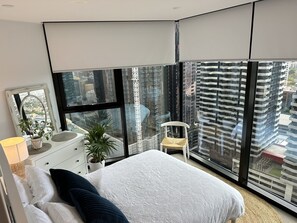 A modern bedroom with a large bed, a dresser, a chair, and a view of a cityscape through large windows.