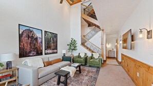 A spacious living room with a sofa, coffee table, and green armchairs. A staircase leads to an upper level. - Luxe Chalet - Sleeps 15 (Midway)