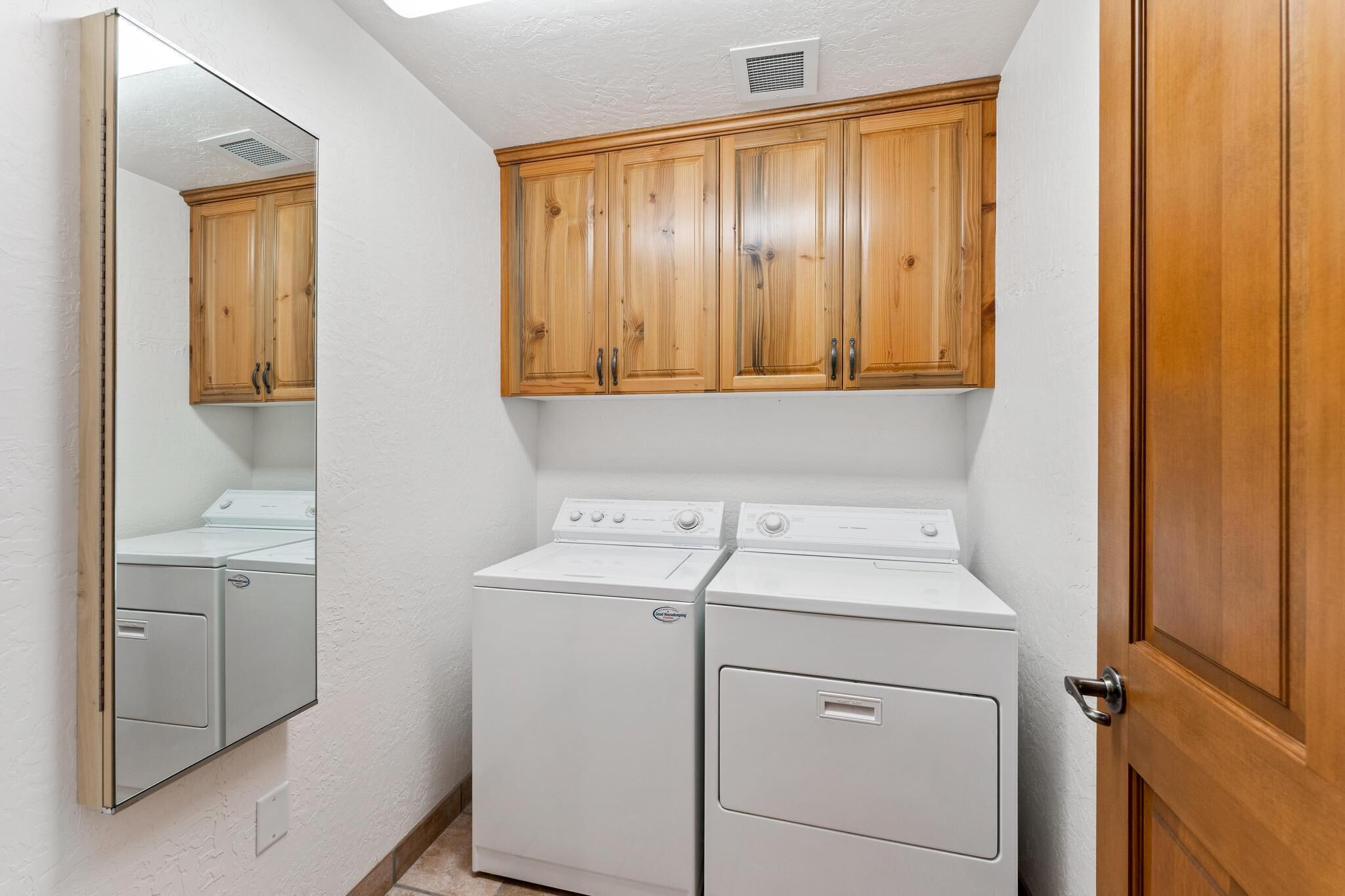 A laundry room with two white front-loading washers and dryers, wooden cabinets above, and a mirror on the left wall.