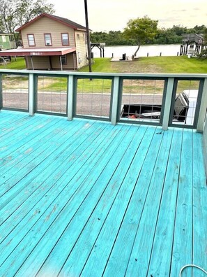 Een turquoise terras met een metalen hekwerk, met uitzicht op een wateroppervlak en een huis op de achtergrond.
