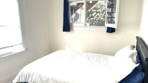 Una cama individual en una habitación con ventana, paredes blancas y vistas a un edificio.