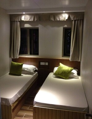 Ein kleiner, schlicht beleuchteter Raum mit zwei Einzelbetten, jeweils mit Kopfstütze und Kissen. Ein Fenster mit Vorhängen und ein kleines Regal mit zwei Steckdosen.