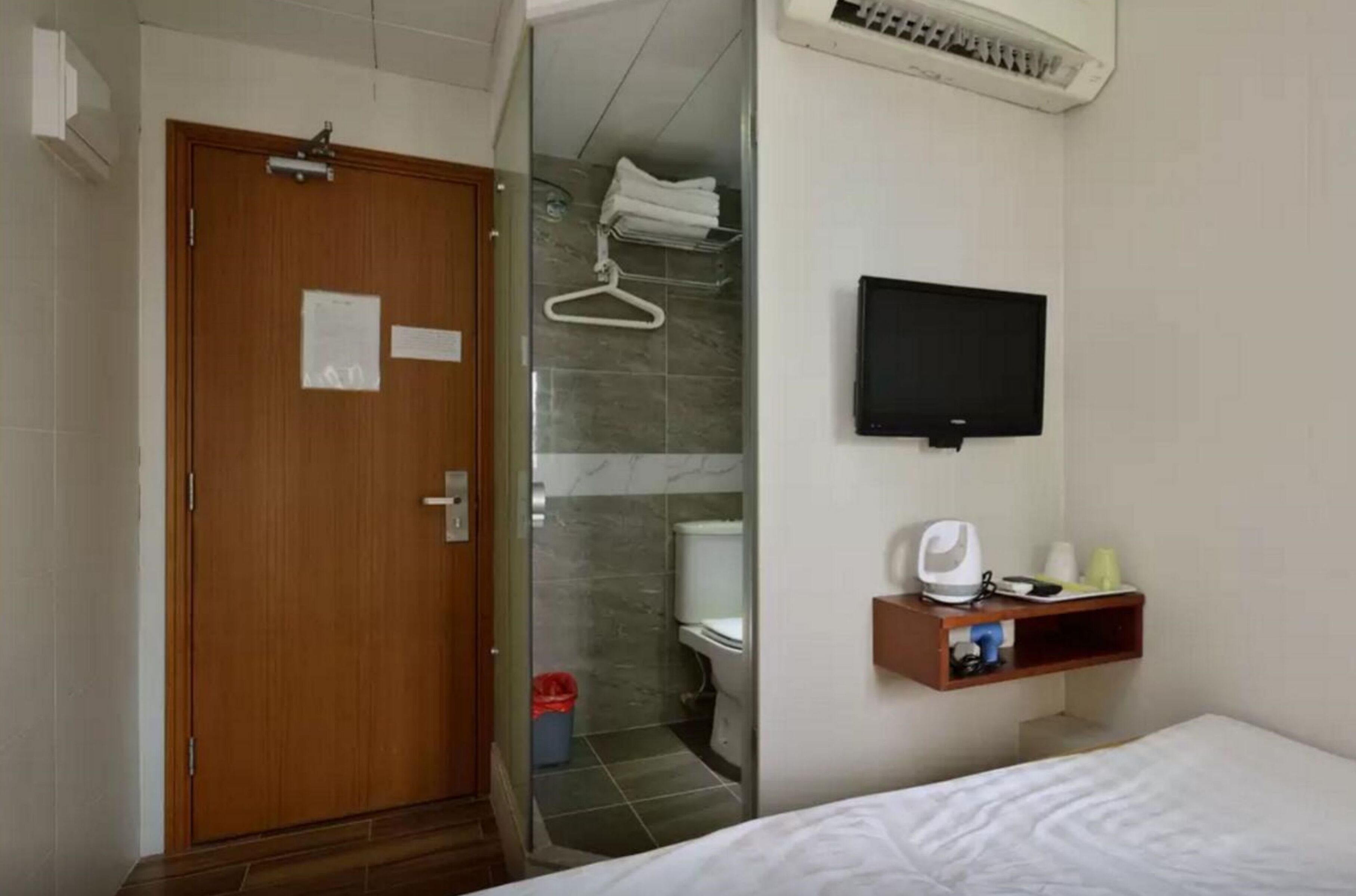 Habitación de hotel con puerta de madera, zona de ducha, televisor montado y estante pequeño con artículos de aseo.