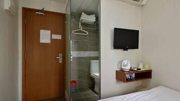 Habitación de hotel con puerta de madera, zona de ducha, televisor montado y estante pequeño con artículos de aseo.