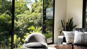 Ein modernes Wohnzimmer mit einem Stehsessel, einem Holztisch und einer Couch mit gestreiften Kissen. Große Fenster bieten Blick auf üppiges Grün.