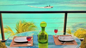 Una mesa redonda de vidrio con dos platos, una botella verde y dos vasos, sobre un balcón con vista al mar turquesa y un barco a lo lejos.