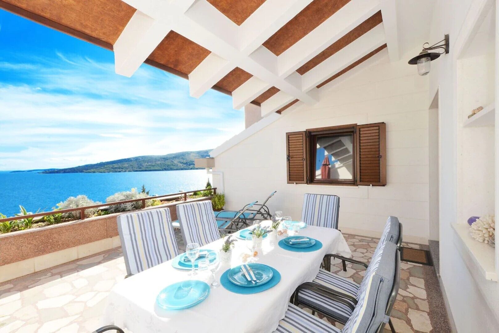 Una mesa preparada para dos personas, con platos y vasos azules, un mantel blanco y vista al mar.
