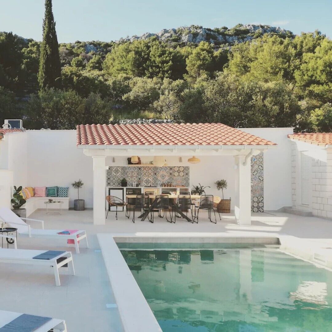 Un área junto a la piscina con pérgola, mobiliario y un muro de azulejos.