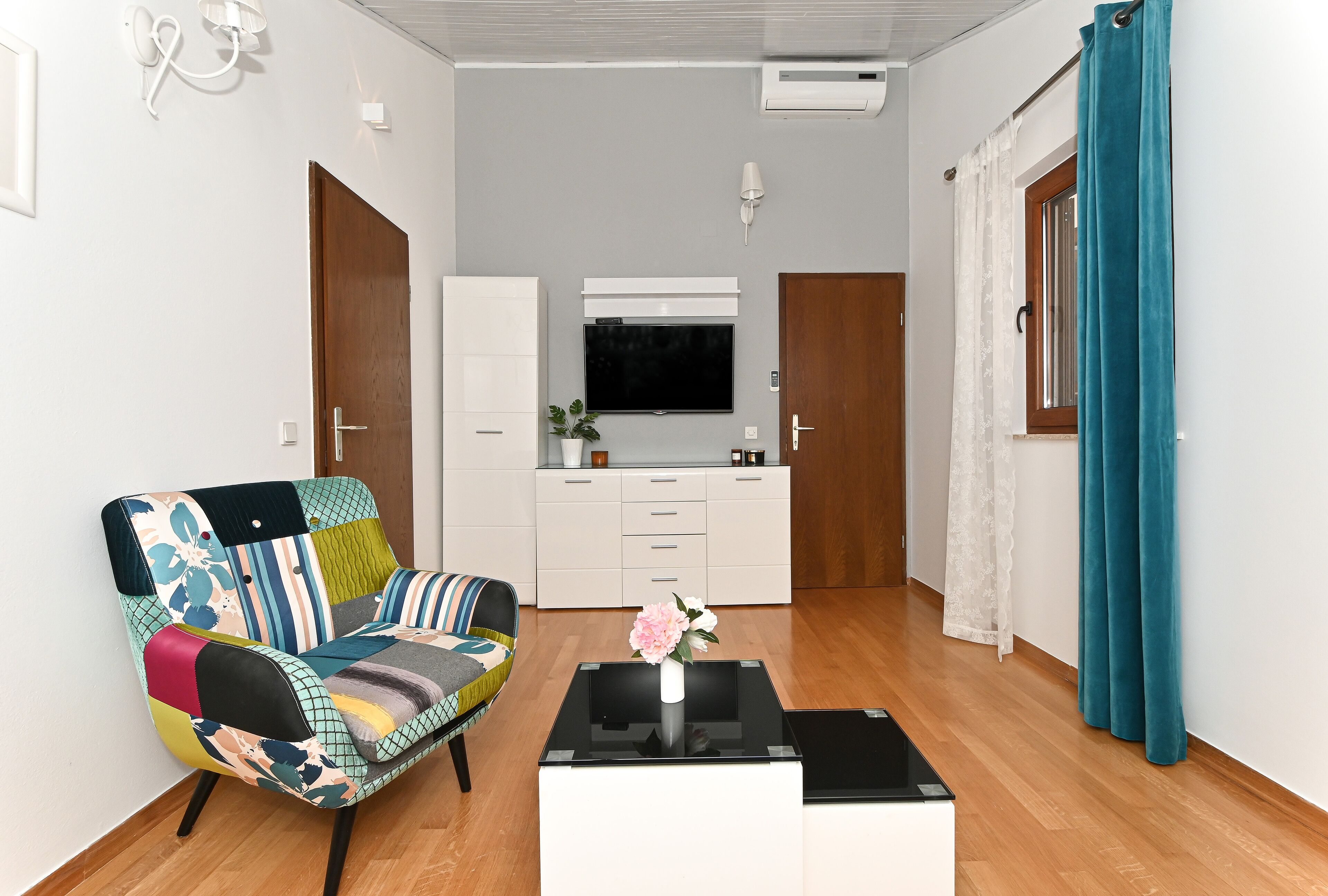 Un salon moderne avec un canapé coloré, une table basse agrémentée d’un vase, un téléviseur à écran plat et un coin cuisine doté de placards et d’une fenêtre.