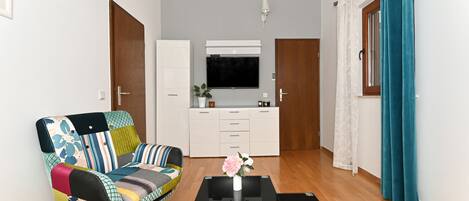 Un salon moderne avec un canapé coloré, une table basse agrémentée d’un vase, un téléviseur à écran plat et un coin cuisine doté de placards et d’une fenêtre.