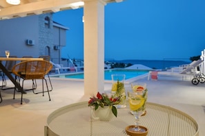 A terrace with a pool, dining area, and a view of the sea at dusk.