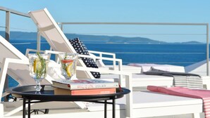 Una terraza iluminada por el sol con sillones de descanso, una mesa con bebidas y libros, y vistas al mar y montañas distantes.