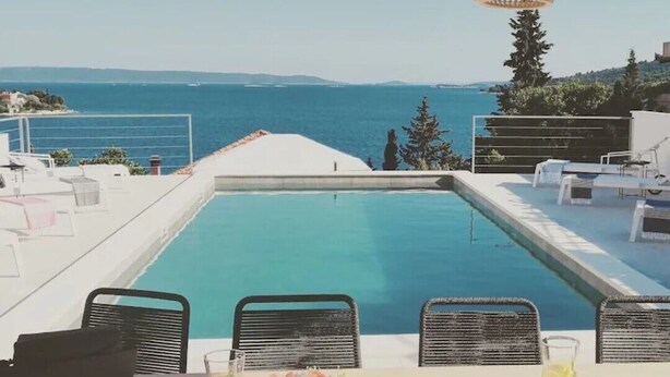 Zona de comedor junto a la piscina con vista al mar, mobiliario exterior y una mesa preparada con frutas y bebidas.