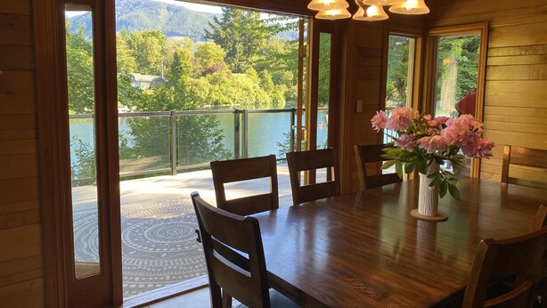 Una mesa de comedor de madera con sillas, un jarrón de flores y vistas a un lago y montañas a través de amplios ventanales.