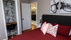 Une chambre avec un lit rouge, un bureau et une chaise dans un coin. On y trouve une porte donnant sur une salle de bain et un couloir.