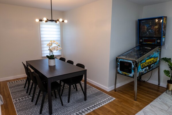 Un coin repas avec une table et des chaises en bois foncé, un tapis à motifs, une orchidée en pot et une borne d’arcade sur le thème de l’eau.