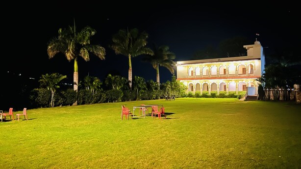A well-lit building with a traditional architectural style, surrounded by a grassy area with palm trees and a few chairs. - PUSHKAR REGENCY RESORT (Pushkar)