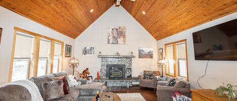 A cozy living room with a fireplace, wooden ceiling, and large windows.