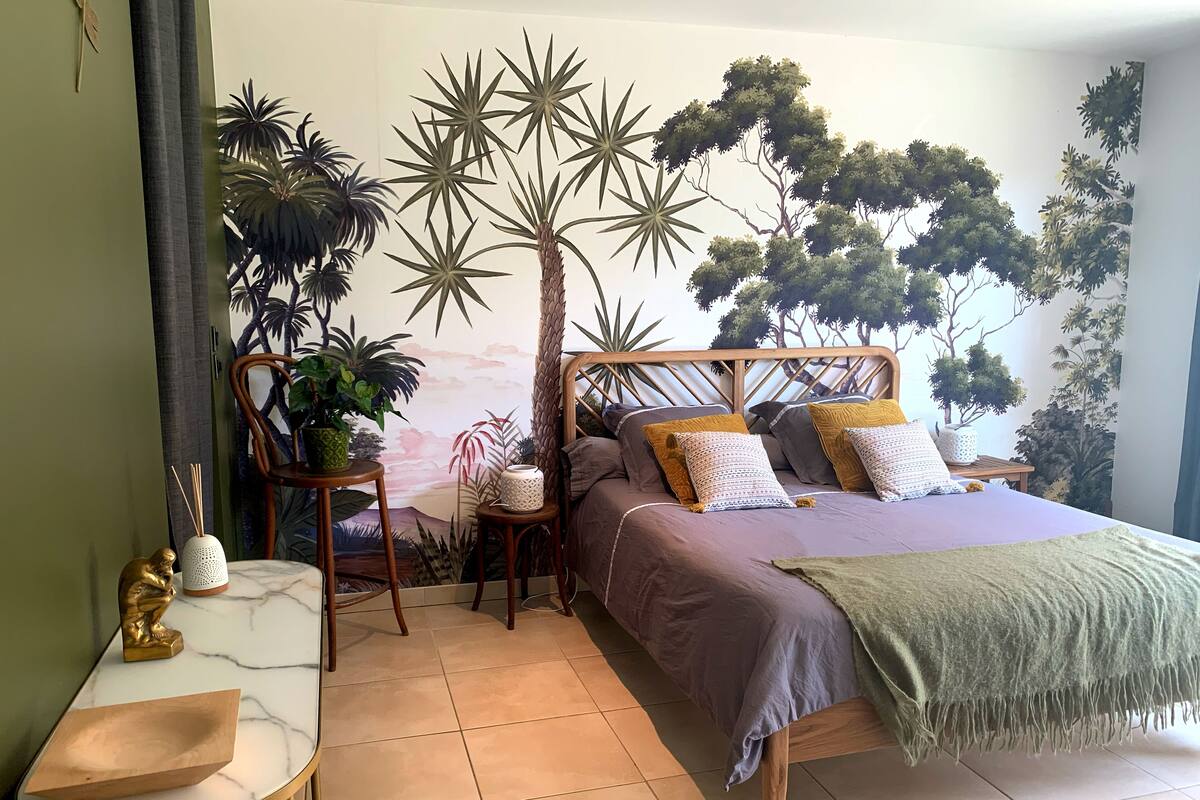 A bedroom with a bed, a chair, a small table, and a wall mural.