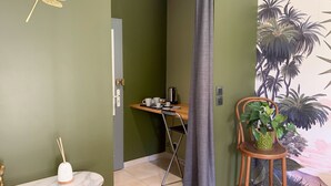 Una habitación con una pared verde, un escritorio de madera con un hervidor, una mesita con un florero blanco y una planta en maceta.