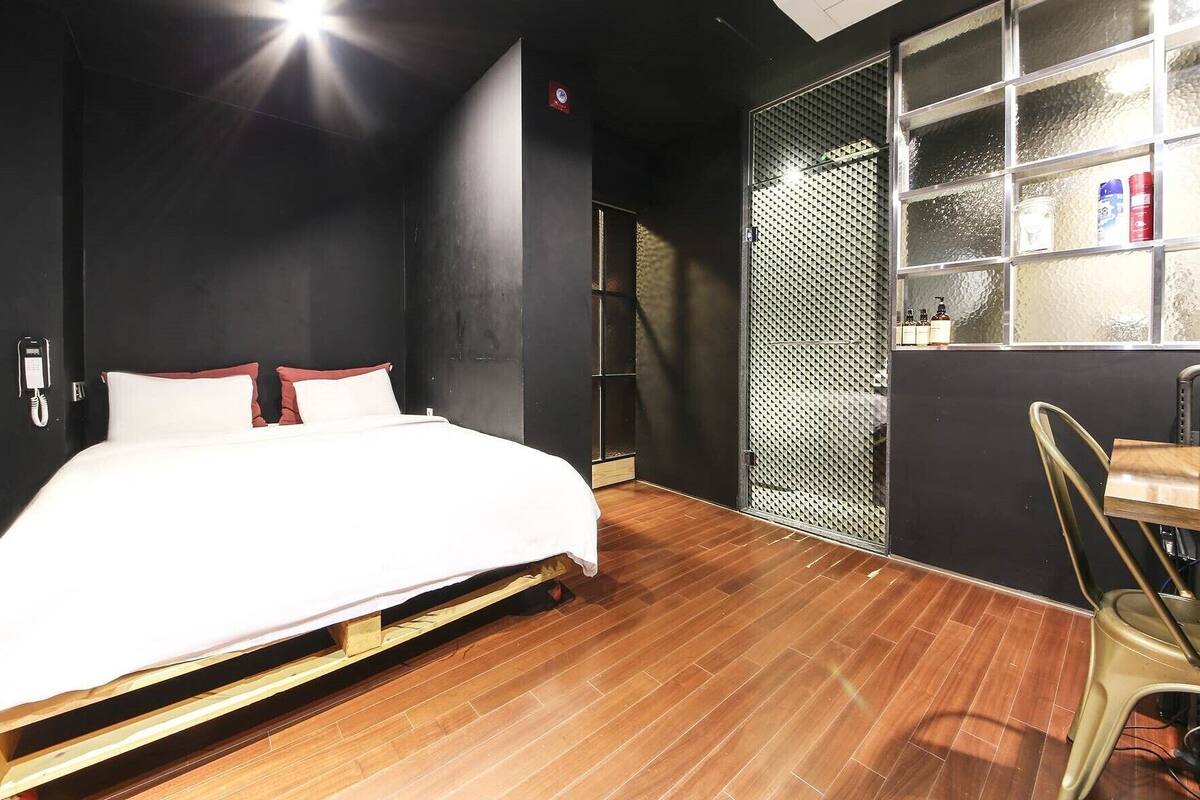 A hotel room with a bed, wooden flooring, a small table, and a window with blinds.