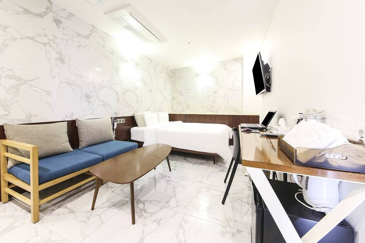 A hotel room with a bed, a sofa, a desk with a laptop, and a TV mounted on the wall.