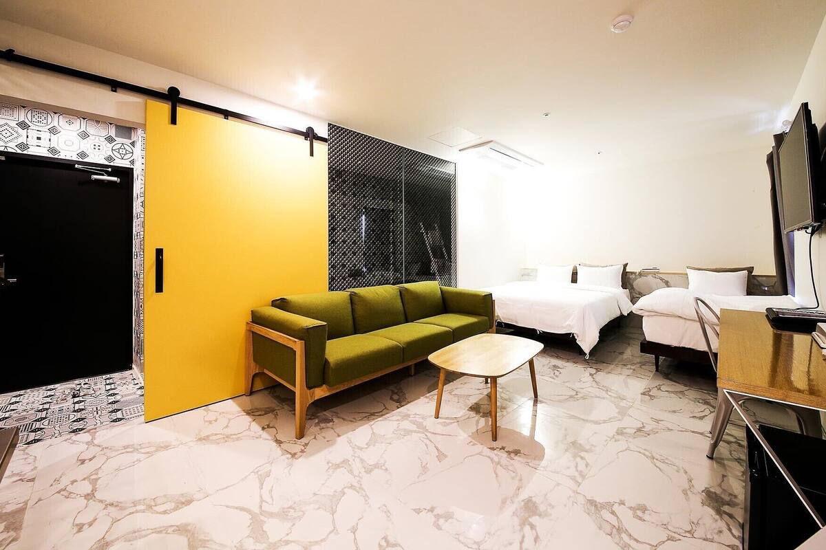 A compact hotel room with a green sofa, two single beds, a small table, and a TV.