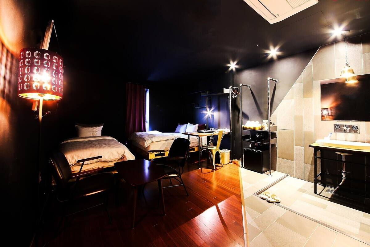 A hotel room with two beds, a desk, a chair, a desk lamp, a TV, and a bathroom with a sink and mirror.