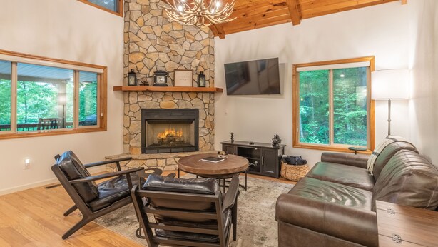 A cozy living room with a stone fireplace, leather furniture, and large windows offering a view of the outdoors.