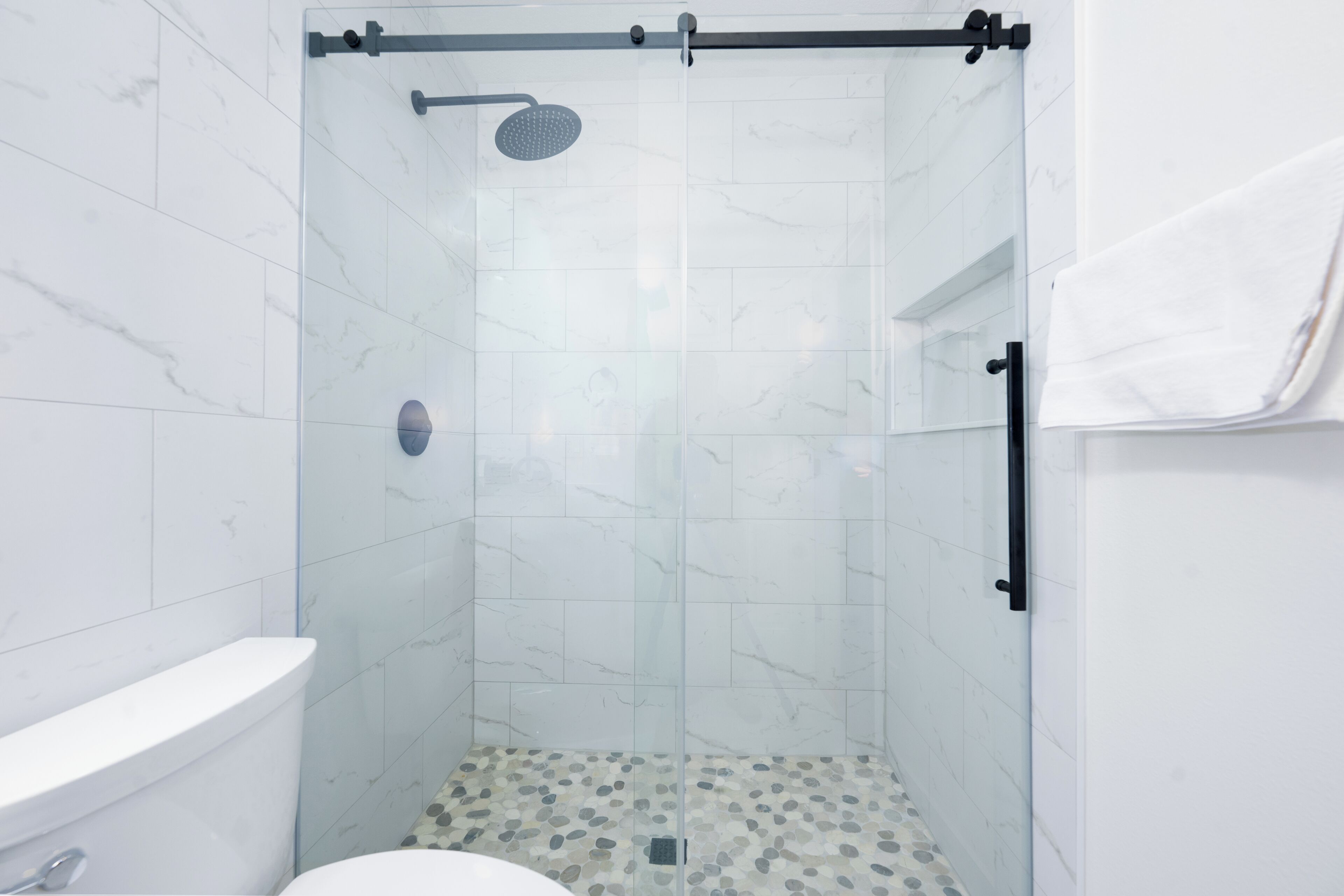 Un baño moderno con ducha de vidrio, baldosas de mármol blanco y piso con diseño de guijarros.