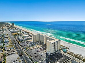 An aerial view of a coastal hotel with a parking lot, a beach, and a clear blue ocean. - Beachfront! Heated pool, spa, Tiki bar & Views! (Panama City Beach)