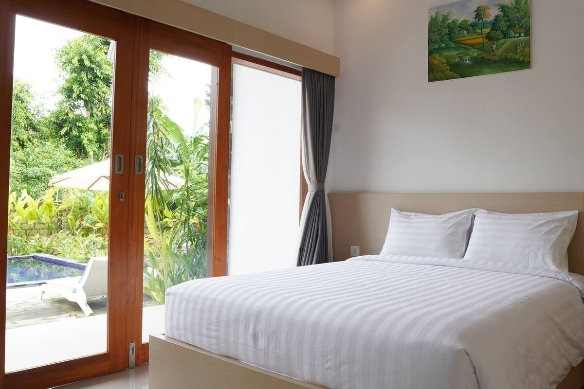 Een hotelkamer met een bed, een schilderij aan de muur en uitzicht op een tuin door de schuifglazen deur.