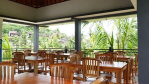 A covered outdoor dining area with wooden tables and chairs, large windows offering views of a lush, green landscape with hills and buildings in the distance. - Artaloka Hotel & Restaurant (Penida Island)