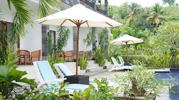 Zona de piscina con sillones de descanso, sombrillas y abundante vegetación.