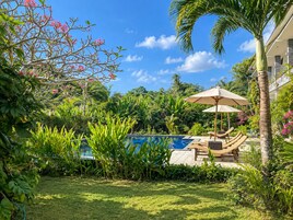 Un área de piscina con sillones de descanso y una sombrilla, rodeada de exuberante vegetación y un cielo despejado.